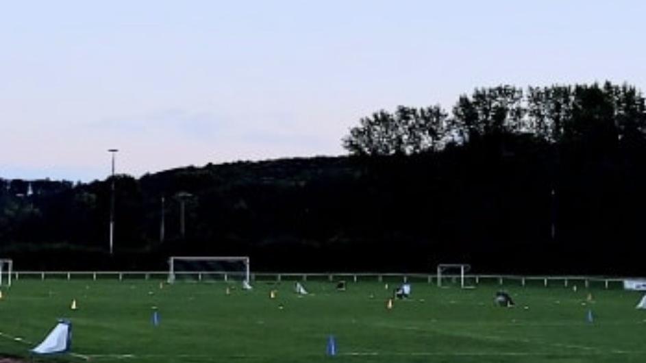 Fu&szlig;balltraining auf einem Rasenplatz mit Markierungen und zwei Toren im Hintergrund. Banner mit der Aufschrift &bdquo;indula&ldquo;.