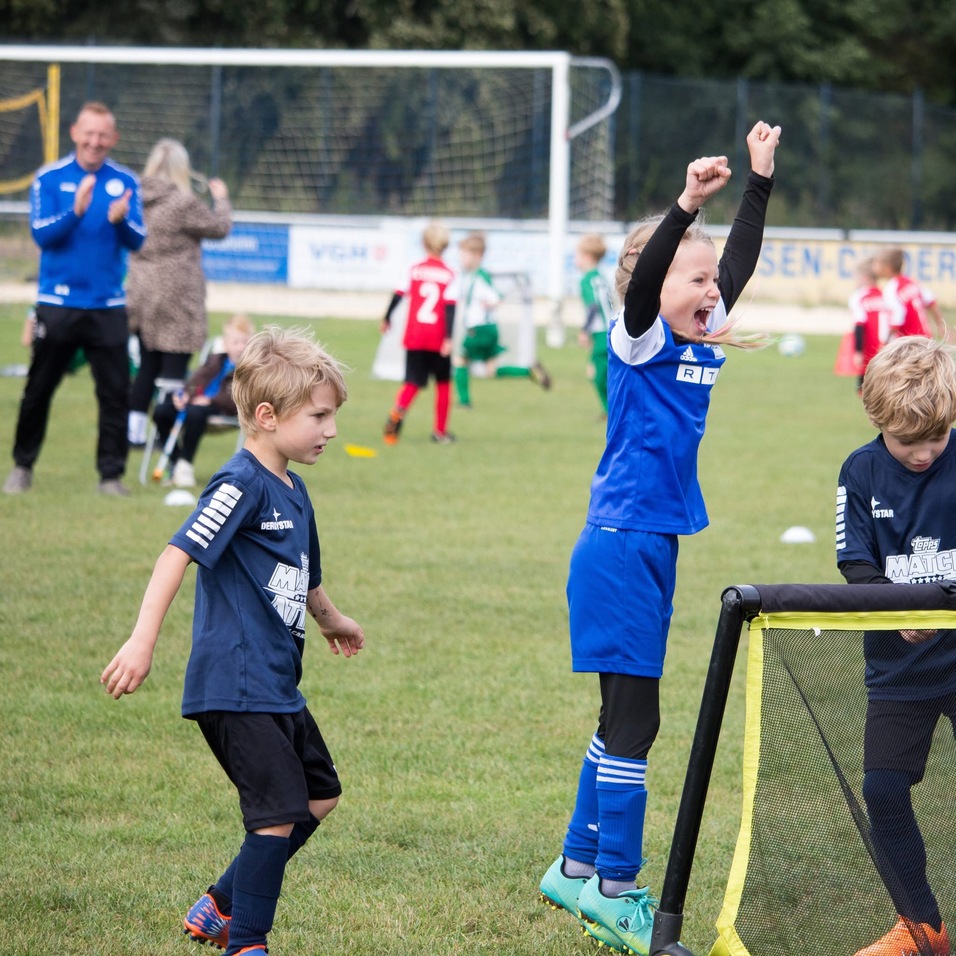 Zwei Kinder jubeln begeistert auf einem Fu&szlig;ballplatz, w&auml;hrend ein drittes Kind neben ihnen steht und zuschaut.