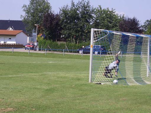 Torwart kniet im Torraum, w&auml;hrend ein Fu&szlig;ball vor ihm auf dem Rasen liegt, im Hintergrund stehen B&auml;ume und Autos.