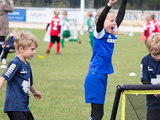 Zwei Kinder jubeln begeistert auf einem Fu&szlig;ballplatz, w&auml;hrend ein drittes Kind neben ihnen steht und zuschaut.