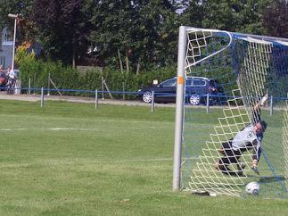 Torwart kniet im Torraum, während ein Fußball vor ihm auf dem Rasen liegt, im Hintergrund stehen Bäume und Autos.