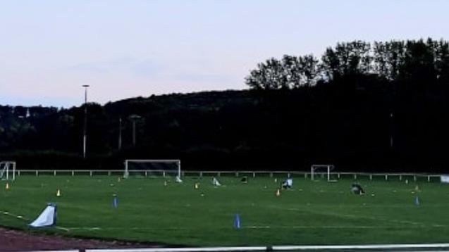 Fußballtraining auf einem Rasenplatz mit Markierungen und zwei Toren im Hintergrund. Banner mit der Aufschrift „indula“.
