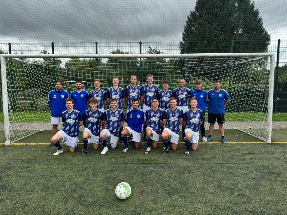 Gruppenshot eines Fu&szlig;ballteams in blauen Trikots vor einem Tor auf einem Kunstrasenplatz. Ein Ball liegt im Vordergrund.