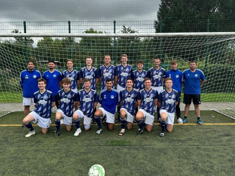 Gruppenshot eines Fu&szlig;ballteams in blauen Trikots vor einem Tor auf einem Kunstrasenplatz. Ein Ball liegt im Vordergrund.