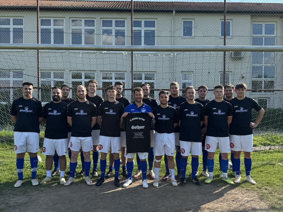 Mannschaftsaufstellung von 14 Fu&szlig;ballspielern in schwarzen Trikots vor einem Tor auf einem Sportplatz.