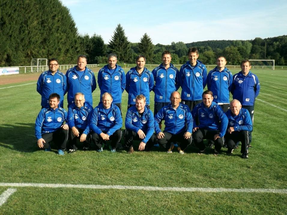 Eine Gruppe von 15 Trainern und Betreuern in blauen Jacken auf einem Fu&szlig;ballplatz vor B&auml;umen und einem klaren Himmel.
