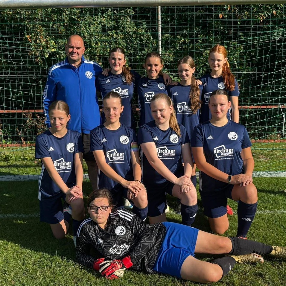 Mannschaftsfoto einer Fu&szlig;ball-Damenauswahl in blauen Trikots, auf einem Spielfeld mit Tor im Hintergrund.