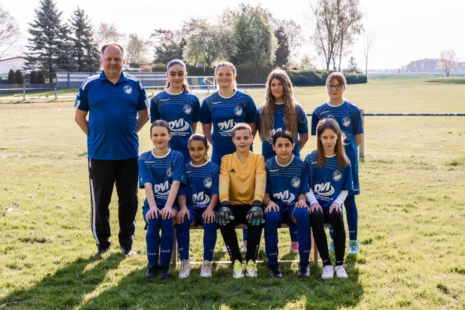 Teamfoto einer M&auml;dchenfu&szlig;ballmannschaft in blauen Trikots mit Trainer auf einem gr&uuml;nen Spielfeld.