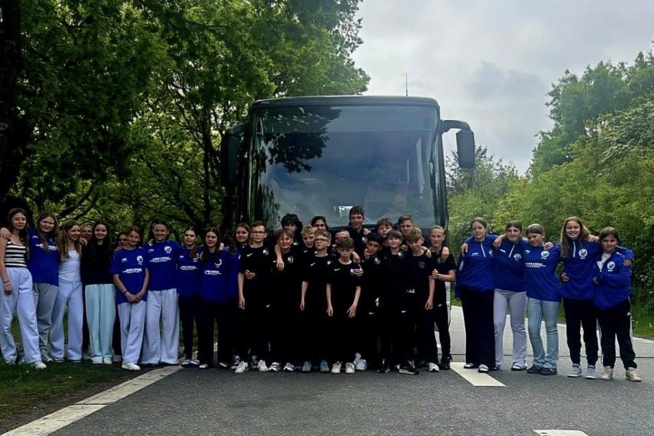 Gruppe junger Sportlerinnen in einheitlicher Kleidung posiert vor einem Bus auf einer Stra&szlig;e, umgeben von B&auml;umen.