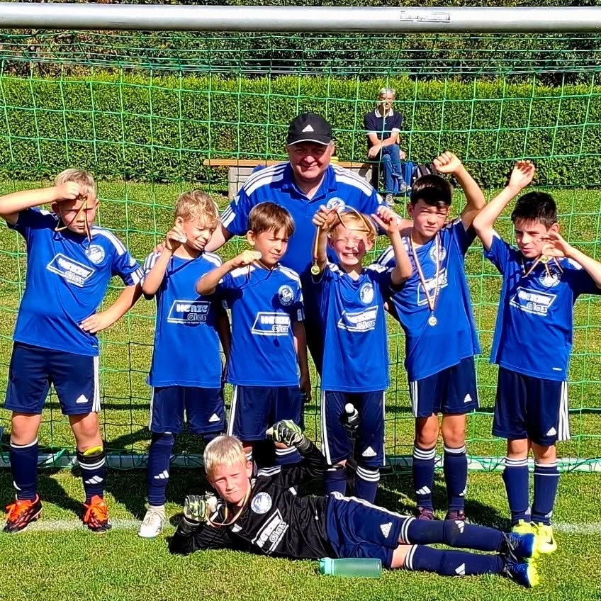 Gruppe von sieben jungen Fu&szlig;ballspielern in blauen Trikots posieren mit Medaillen vor einem Fu&szlig;balltor.