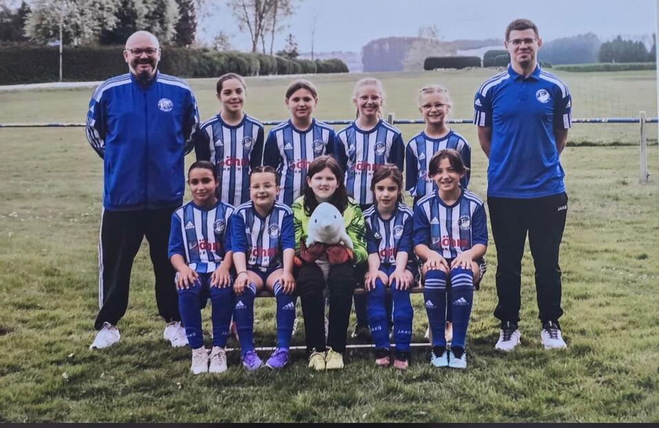 Mannschaftsfoto einer Fu&szlig;ball-Jugendmannschaft mit Trainer auf Rasenplatz, in blauen Trikots und einheitlichem Outfit.