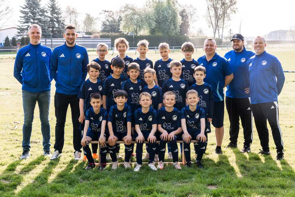 Gruppenfoto einer Fussballmannschaft mit Trainern, alle tragen blaue Trikots und stehen auf einem gr&uuml;nen Spielfeld.