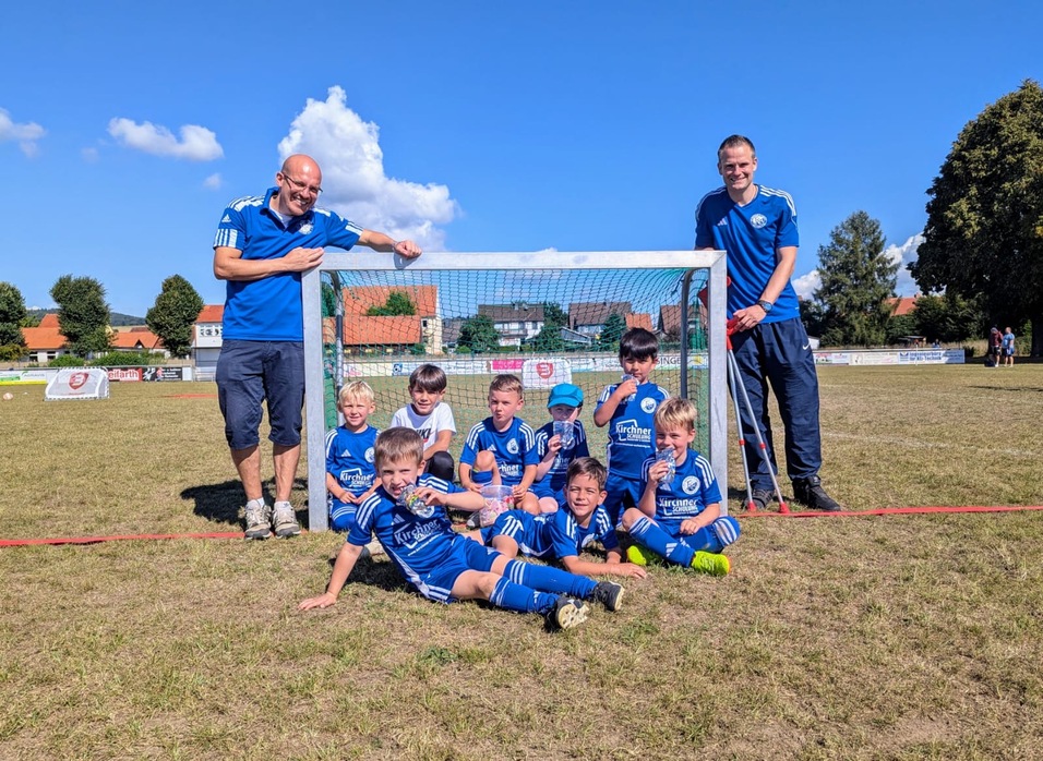 Gruppe von Kindern in blauen Fu&szlig;balltrikots posiert vor einem Fu&szlig;balltor auf einem Rasenplatz, umrahmt von B&auml;umen.