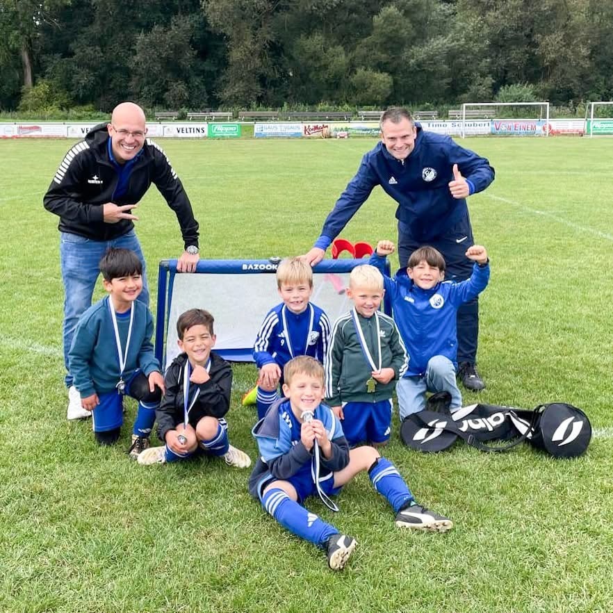 Gruppe von sechs jubelnden Kindern in Fu&szlig;balltrikots mit Medaillen, posierend vor einem Tor, zusammen mit zwei Trainern.