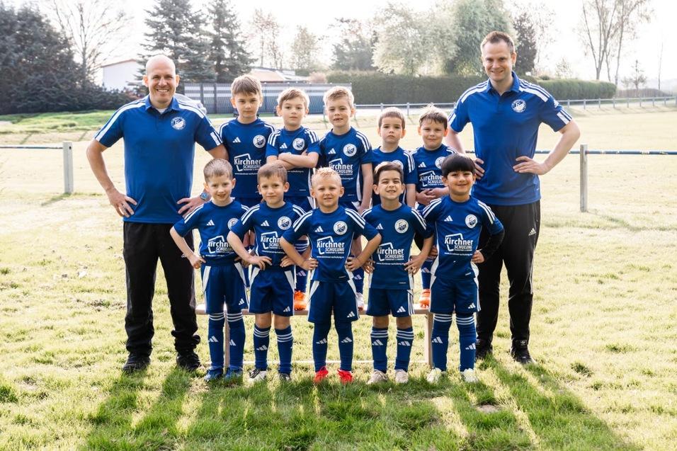 Kinderfu&szlig;ballmannschaft in blauen Trikots, stehen auf einem gr&uuml;nen Rasenplatz, flankiert von zwei Trainern.