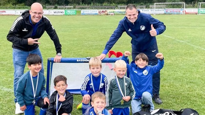 Gruppe von sechs jubelnden Kindern in Fu&szlig;balltrikots mit Medaillen, posierend vor einem Tor, zusammen mit zwei Trainern.