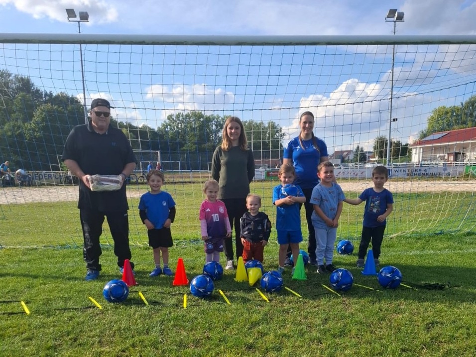 Gruppe von Kindern in Sportkleidung steht mit zwei Erwachsenen vor einem Fu&szlig;balltor, umgeben von B&auml;llen und H&uuml;tchen auf dem Rasen.