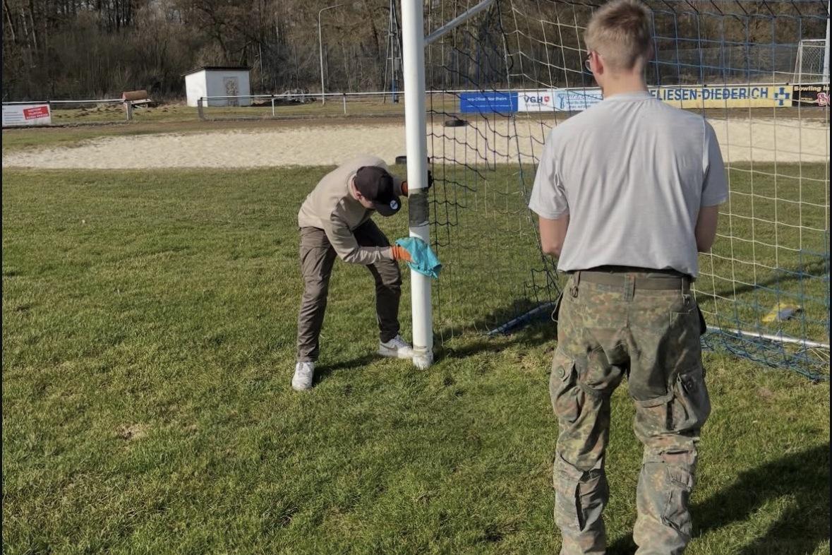 Zwei Personen befestigen ein Netz an einem Fu&szlig;balltor auf einem Sportplatz bei sonnigem Wetter.