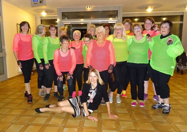 Gruppe von Frauen in bunten Netzshirts posiert gemeinsam in einer Halle, eine Frau sitzt vorne im Vordergrund.