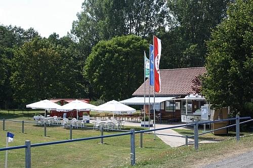 Blick auf ein Freizeitgelände mit einem Kiosk, Tischen, Sonnenschirmen und Fahnen vor einer grünen Landschaft.