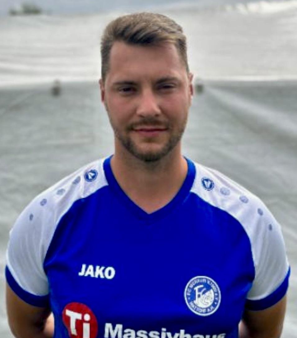 Portrait eines Mannes in einem blauen Fu&szlig;balltrikot mit Logos, vor einem grauen Hintergrund.