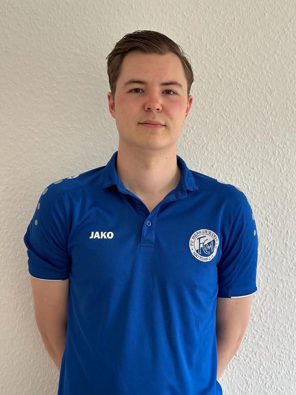 Junger Mann in blauem Sporttrikot mit Logo steht vor einer hellen Wand.