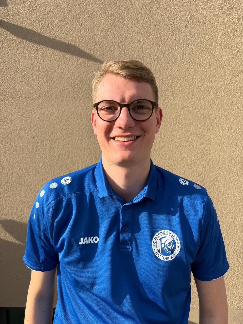 L&auml;chelnder Mann in blauer Sportkleidung mit Brille steht vor einer hellen Wand.