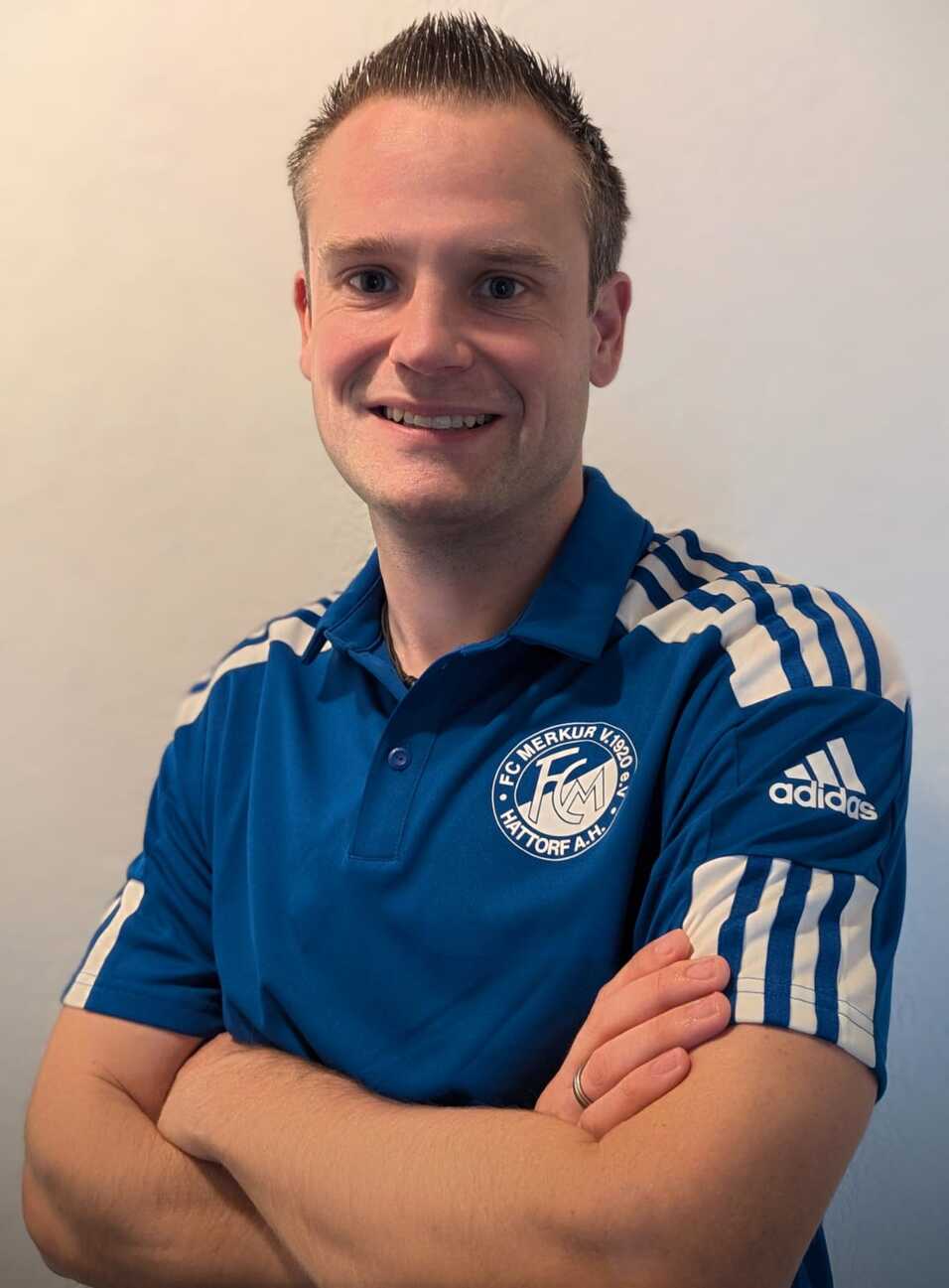 L&auml;chelnder Mann in blauer Sportbekleidung mit wei&szlig;en Streifen und Vereinslogo steht mit verschr&auml;nkten Armen vor einer neutralen Wand.