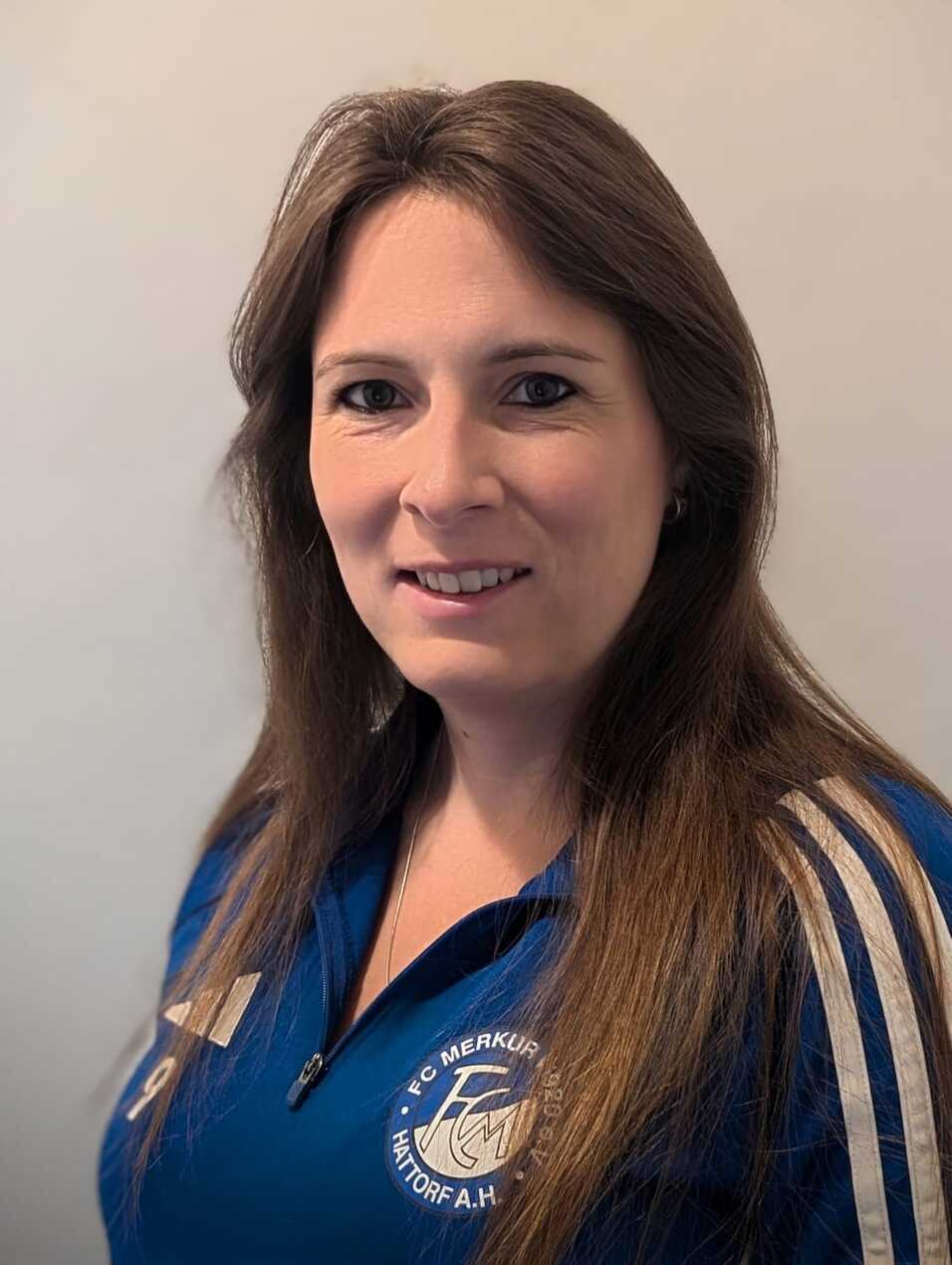 Portrait einer Frau mit braunen Haaren in blauer Sportjacke des FC Merkur Hattorf, l&auml;chelnd vor einem hellen Hintergrund.