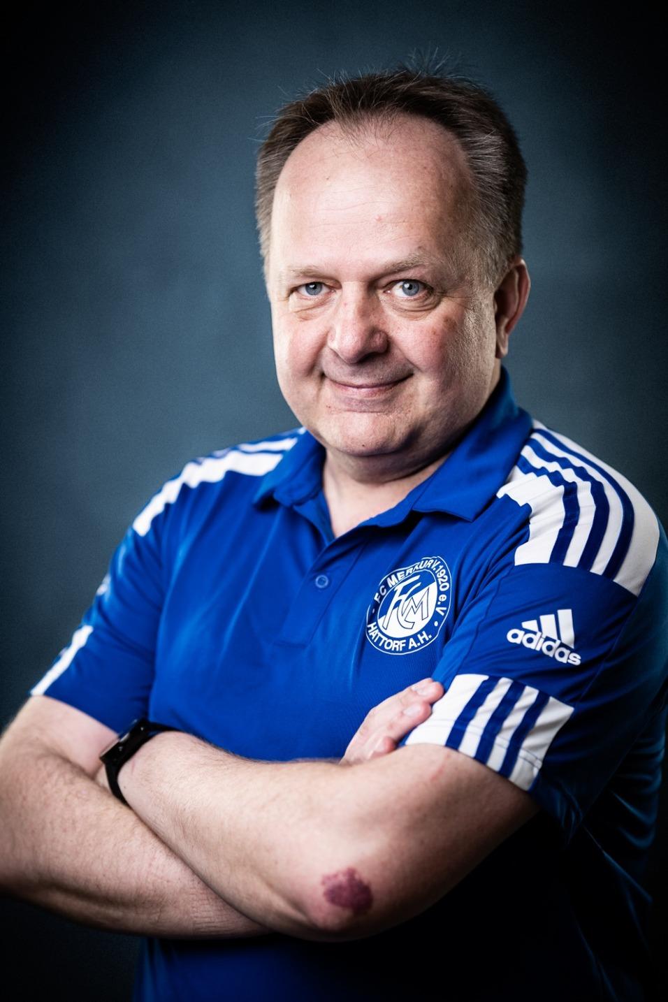 Portr&auml;t eines l&auml;chelnden Mannes in blauer Sportbekleidung mit Armen vor der Brust verschr&auml;nkt auf dunklem Hintergrund.