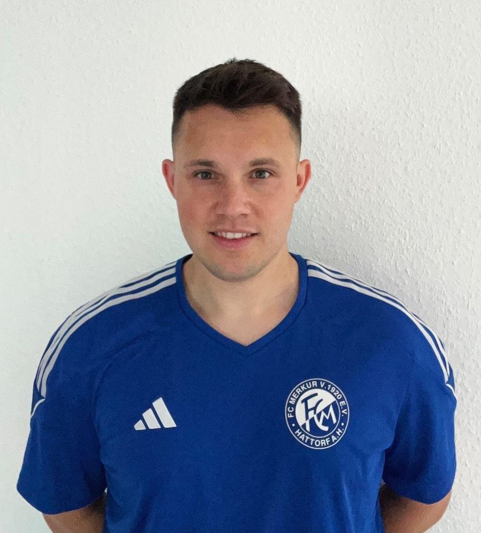 Mann in blauem Sporttrikot mit Vereinslogo steht vor einer wei&szlig;en Wand und l&auml;chelt in die Kamera.