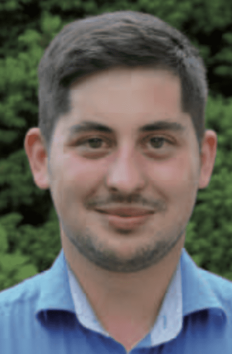 Portr&auml;t eines jungen Mannes mit kurzem, dunklem Haar, der in einem blauen Hemd vor gr&uuml;ner Hintergrundvegetation l&auml;chelt.